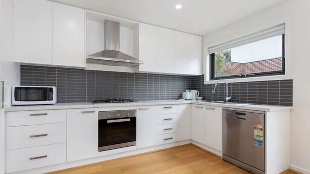 A modern kitchen with white cabinetry, a stainless steel range hood, a microwave, an oven, and a dishwasher. - Domi Rentals - The Mill Residence (Clayton South)