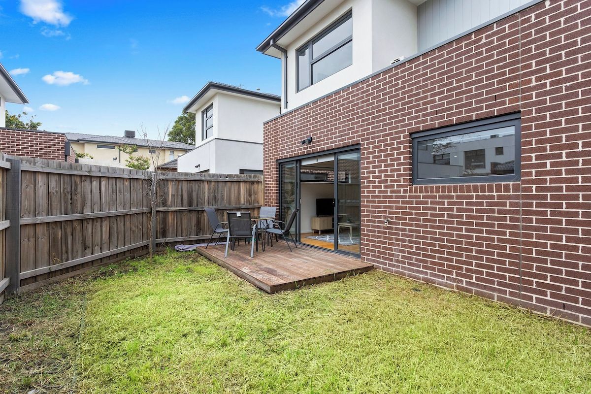A residential backyard with a wooden deck, a brick wall, and a glass door leading to a patio area.
