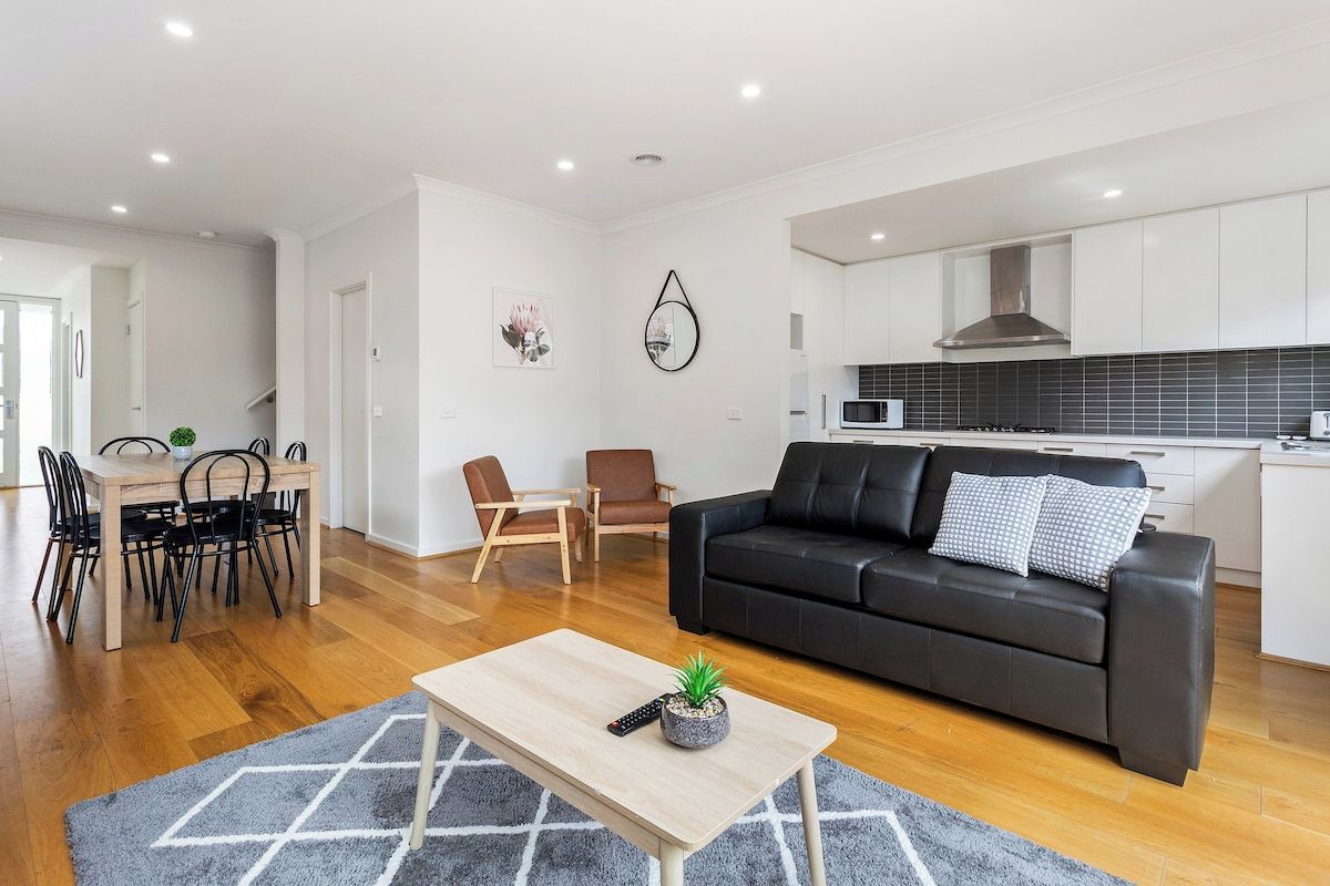 A modern living room with a black leather sofa, a wooden coffee table, and a dining area with a wooden table and chairs.