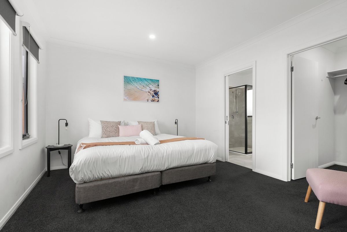 A bedroom with a bed, bedside tables, a bench, a painting, and a glass door leading to a bathroom.