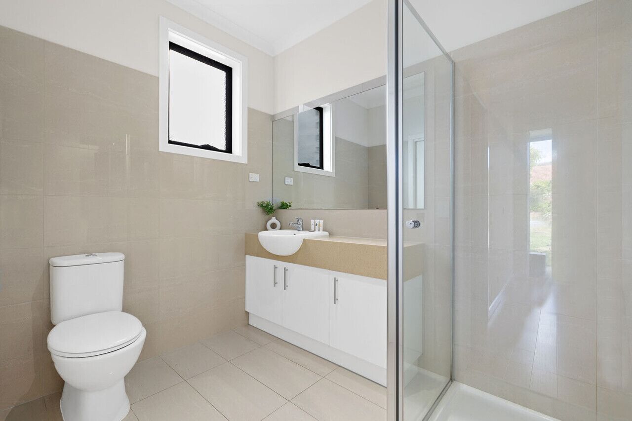 A modern bathroom with a toilet, a sink, a mirror, and a glass shower enclosure. - Domi Rentals - The Fregon Townhouse (Clayton)