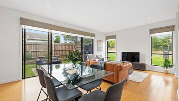 A modern living room with a glass dining table, comfortable seating, and large windows offering a view of the backyard.
