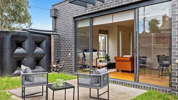 A modern house with a patio area featuring two chairs and a table, a large sliding glass door leading to an interior space with a sofa, and a brick exterior.