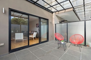 A modern patio with a glass door leading to an interior space, two red chairs, and a small table with a plant. - Domi Rentals - The Chadstone Homes (Chadstone)