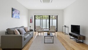 A modern living room with a grey sofa, a coffee table, a TV stand, and a large window with blinds. - Domi Rentals - The Atkinson Home (Chadstone)