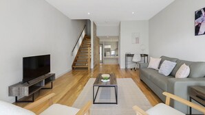 A modern living room with a flat-screen TV, wooden coffee table, grey sofa, and a staircase leading to another room. - Domi Rentals - The Atkinson Home (Chadstone)