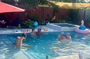 A swimming pool with people enjoying the water, surrounded by a wooden fence, a red umbrella, and various poolside items. - Men Only Heated Pool Spa - Kings Cove @Blue Lizard (Oakland Park)