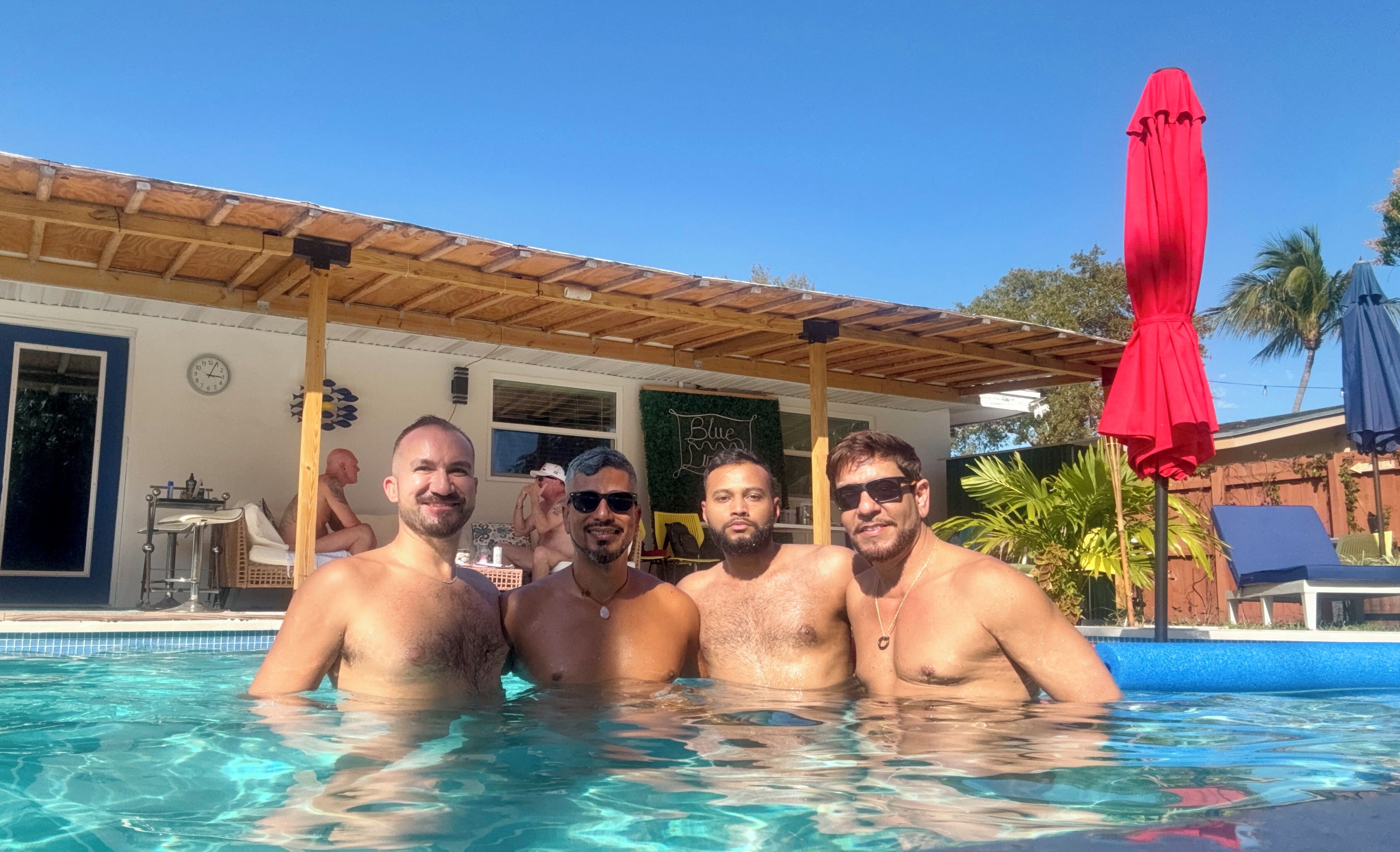Una piscina con quattro persone, una zona coperta con un ombrellone rosso e un edificio con una sveglia e un ornamento a parete.