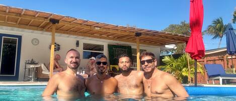 Piscina com quatro pessoas, área coberta com um guarda-sol vermelho e um edifício com relógio e decoração na parede.