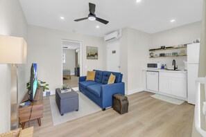 A modern living room with a blue sofa, a coffee table, and a television. There is a kitchen area with a microwave and a dishwasher. - Blue Lizard Men-Only 1/1 Unit with Heated Saltwater Pool and Tropical Backyard (Oakland Park)