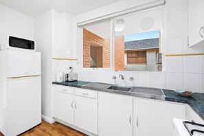 A modern kitchen with white cabinetry, a built-in microwave, a stainless steel sink, and a view of the outside through a window. - Domi Rentals - The Balaclava Den (Saint Kilda East)