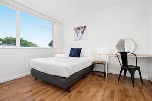 A bedroom with a bed, bedside tables, a desk, a chair, and a window. - Domi Rentals - The Balaclava Den (Saint Kilda East)