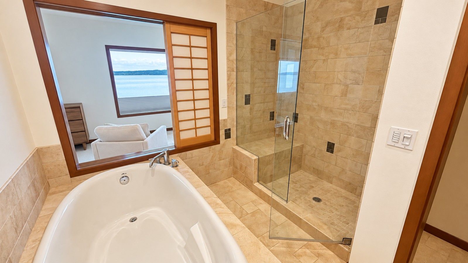 A bathroom with a bathtub, a glass-enclosed shower, and a large mirror reflecting a room with a view of the sea.