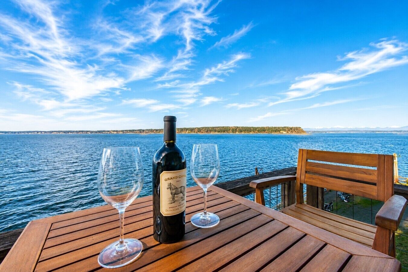 A wooden table with a bottle of wine and two glasses on it, overlooking a body of water with a distant shoreline.