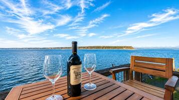 A wooden table with a bottle of wine and two glasses on it, overlooking a body of water with a distant shoreline.