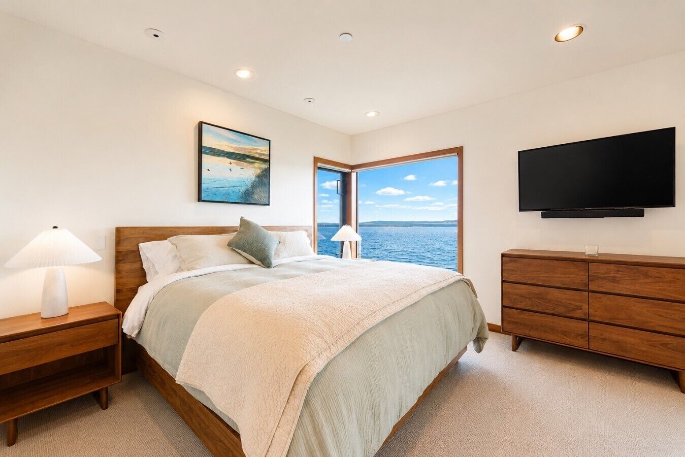 A bedroom with a bed, a nightstand, a dresser, a television, and a window with a view of the sea.