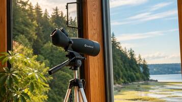 A telescope on a tripod by a window overlooking a forest and water.