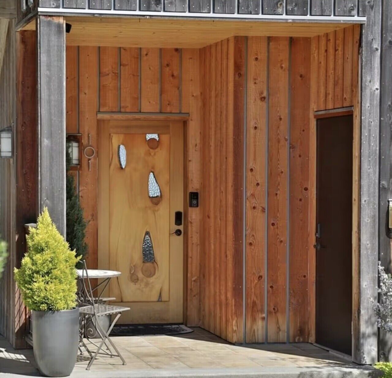 Une porte en bois ornée d’éléments décoratifs, une petite table et une plante en pot devant un bâtiment.