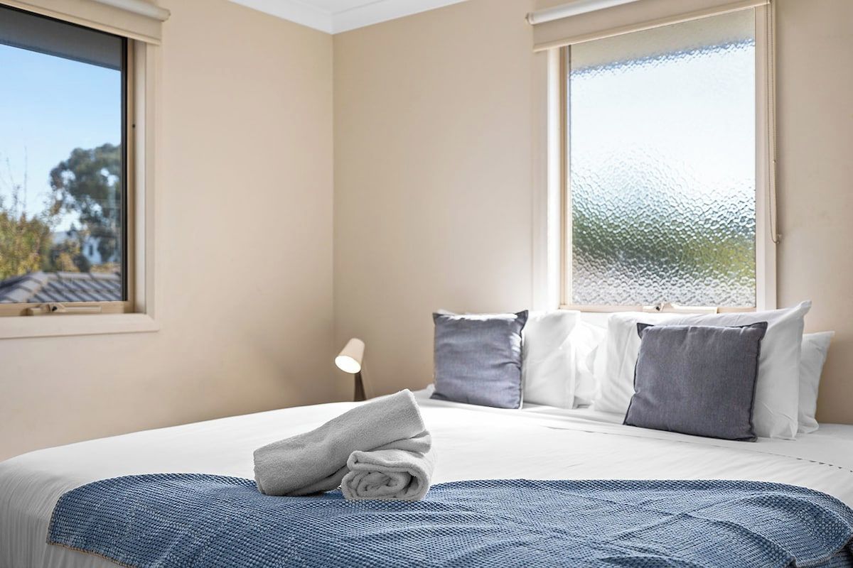 A hotel room with a bed, pillows, a blanket, a window, and a nightstand with a lamp.