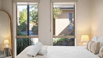 A hotel room with a bed, two windows, a lamp, and a view of a building and greenery outside.