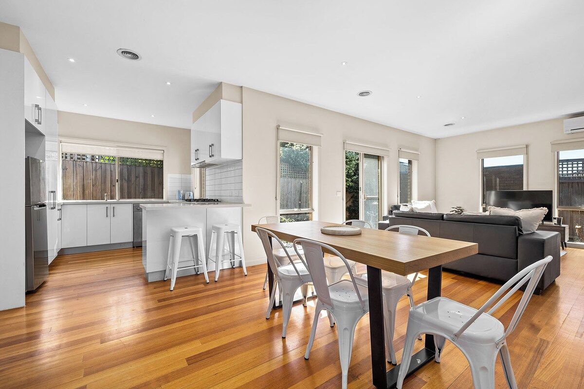 A modern kitchen with white cabinets, a dining area with a wooden table and white chairs, and a living area with a dark sofa and a flat-screen TV.