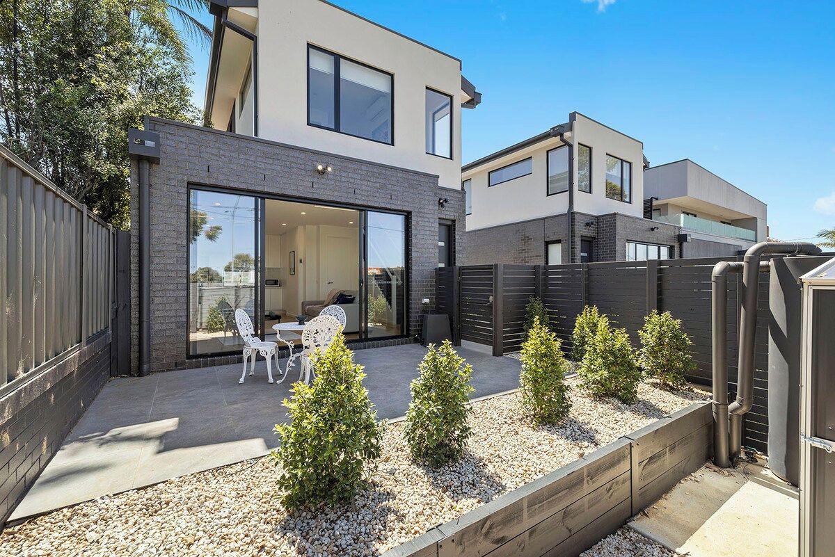 A modern two-story house with a patio area featuring a table and chairs, surrounded by small trees and a wooden fence.