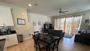 A modern kitchen with white cabinets, a dining area with a dark wooden table and chairs, and a living room with a dark sofa and a large window with curtains. - Condo House 20 min from the Disneyland (Davenport)