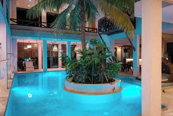 An indoor swimming pool with a central planter, surrounded by a multi-level structure with visible seating areas and a reception desk. - Joanza by OOL Hotels (Isla Holbox)