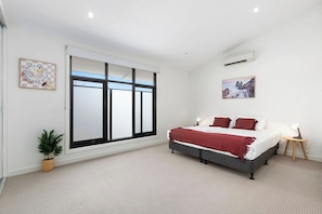 Un dormitorio con cama, ventana, aire acondicionado instalado en la pared y una pequeña mesa con lámpara.