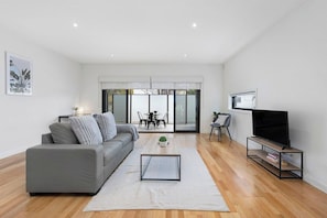 A modern living room with a grey sofa, a coffee table, a flat-screen TV on a stand, and a dining area with a table and chairs. - Domi Rentals - The Chadstone Homes (Chadstone)