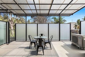 A covered outdoor patio with a table and chairs, a glass partition, and an air conditioning unit. - Domi Rentals - The Chadstone Homes (Chadstone)