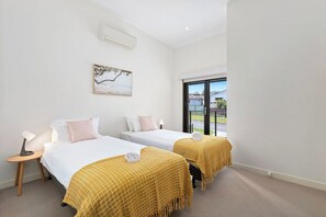A hotel room with two beds, a small table with a lamp, a painting on the wall, and a window with a view of the outside. - Domi Rentals - The Chadstone Homes (Chadstone)