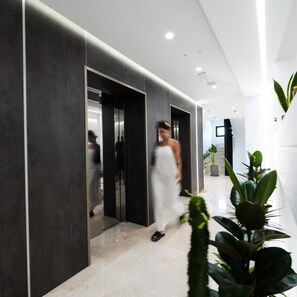 A modern interior with a glass elevator, a person in a white dress, and potted plants. - Naos Luxury Hotel (Orikum)