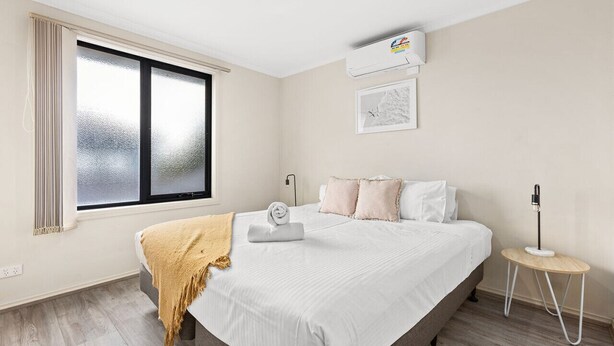 A hotel room with a large bed, a window with curtains, an air conditioning unit, a bedside table with a lamp, and a wall-mounted picture. - Domi Rentals - The Stockdale Home (Clayton)