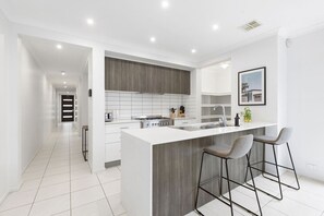 A modern kitchen with a central island, bar stools, and a view of the hallway. - Domi Rentals - The Third Residence (Clayton South)