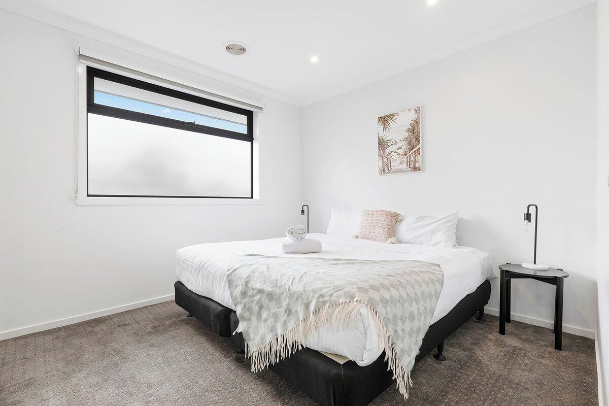 A bedroom with a large bed, a window with blinds, a bedside table, and a wall-mounted artwork.