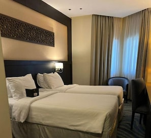 A hotel room with two beds, a desk, a chair, and a large window with curtains.