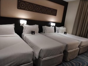 A hotel room with three single beds, each with a pillow and a small bedside table.