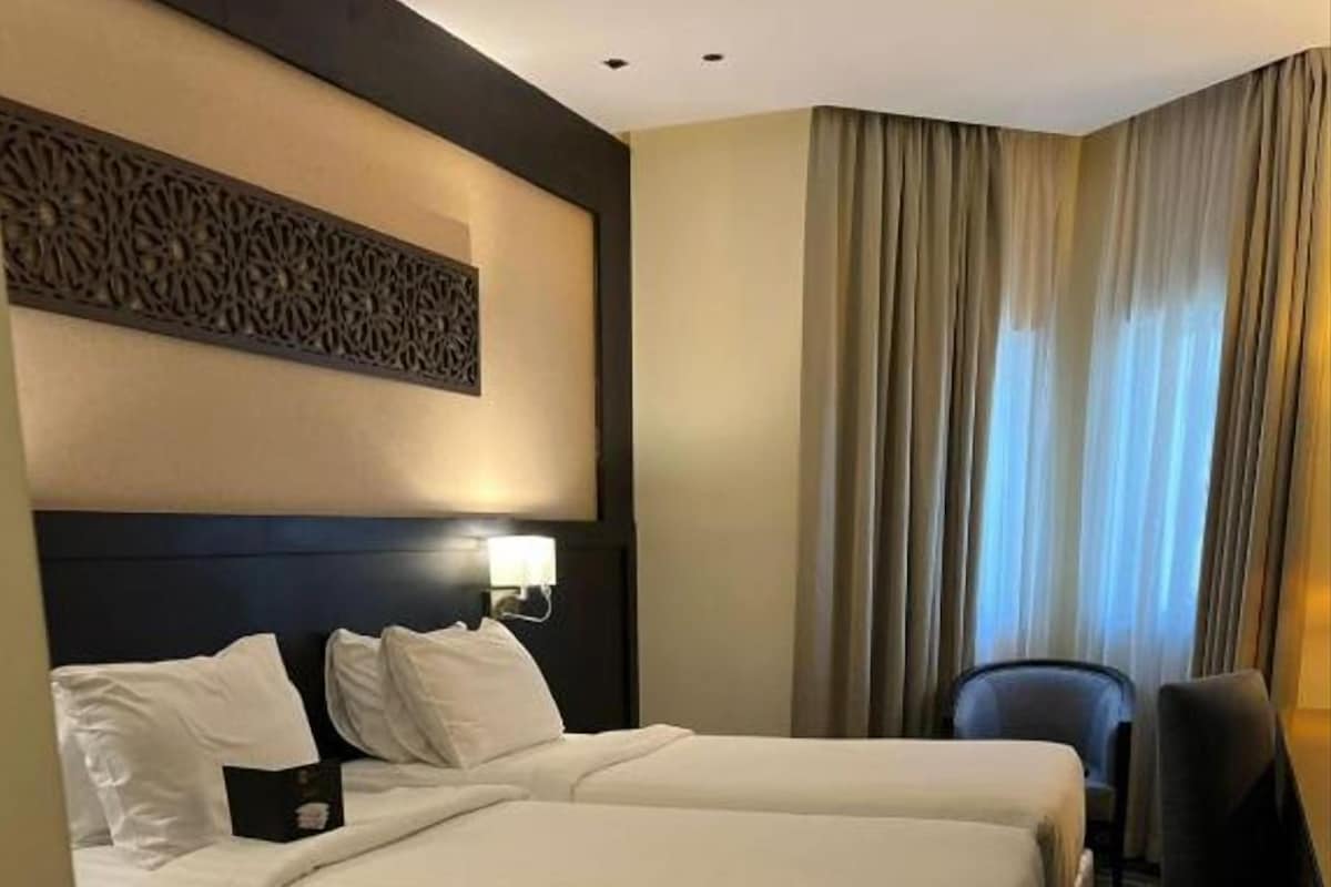 Habitación de hotel con dos camas, un escritorio, una silla y un ventanal con cortinas.