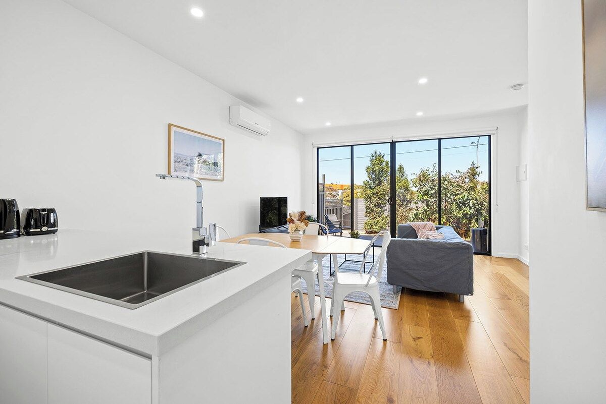 A modern kitchen with a central island, a dining area with a table and chairs, and a living space with a sofa and a large window.