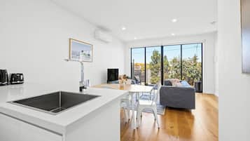 A modern kitchen with a central island, a dining area with a table and chairs, and a living space with a sofa and a large window.