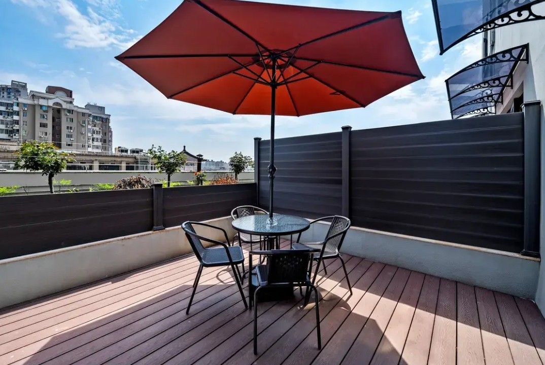 A rooftop terrace with a table and chairs, a red umbrella, and a view of buildings in the background.