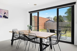 A modern dining area with a wooden table and black chairs, large sliding glass doors leading to a backyard. - Domi Rentals - The Main Residence (Clayton South)