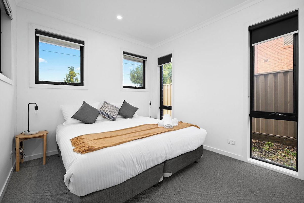 A bedroom with a large bed, two windows, a bedside table, and a view of the outside.