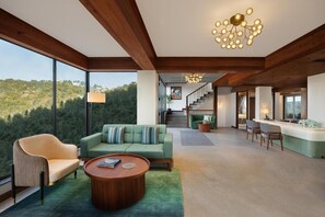 A spacious living area with a large window, a sofa, a coffee table, and a staircase leading to a kitchen area. - Justa Luxe Hollow Oak Mussoorie (Dehradun)