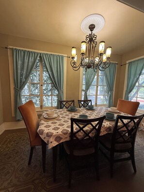A dining area with a table set for four, surrounded by chairs, a chandelier, and large windows with curtains. - Historic House Near Downtown and the Riverwalk (San Antonio)