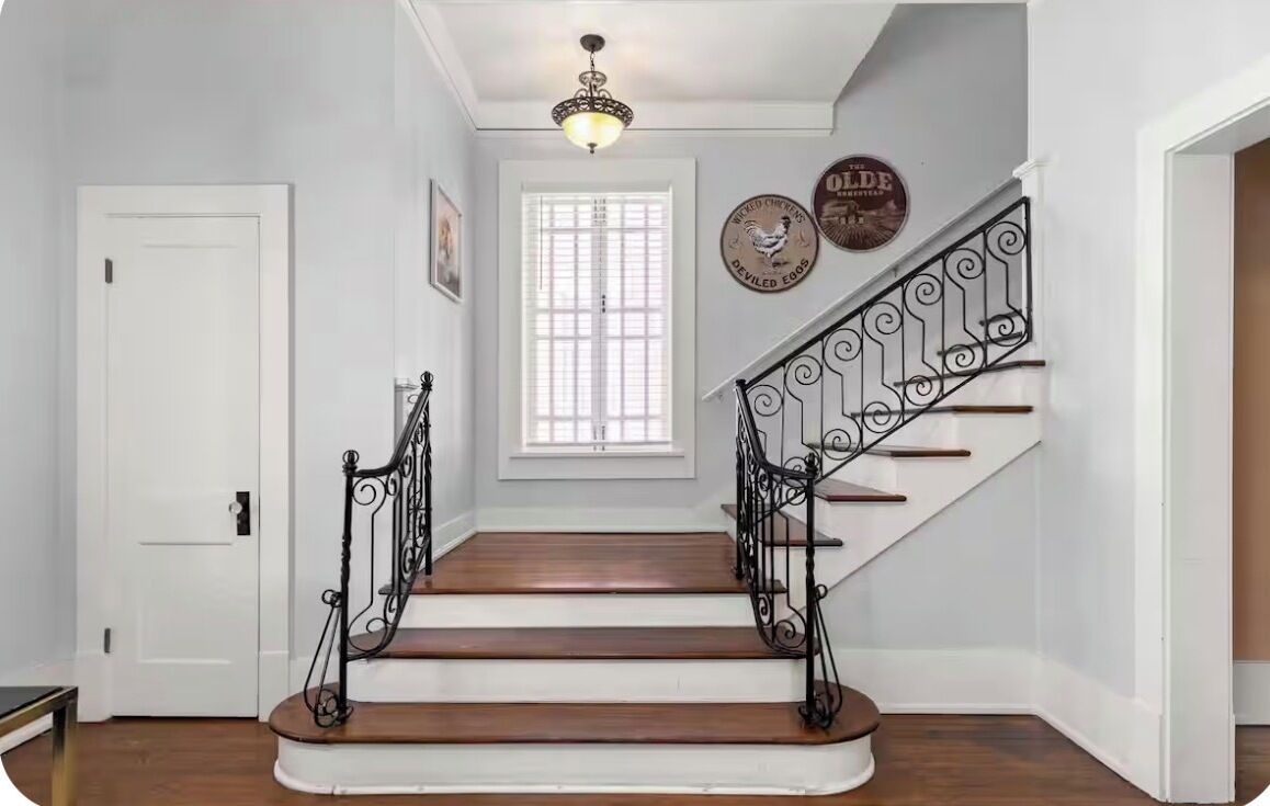 Una escalera con barandillas negras ornamentales, una puerta blanca y un ventanal con persianas.