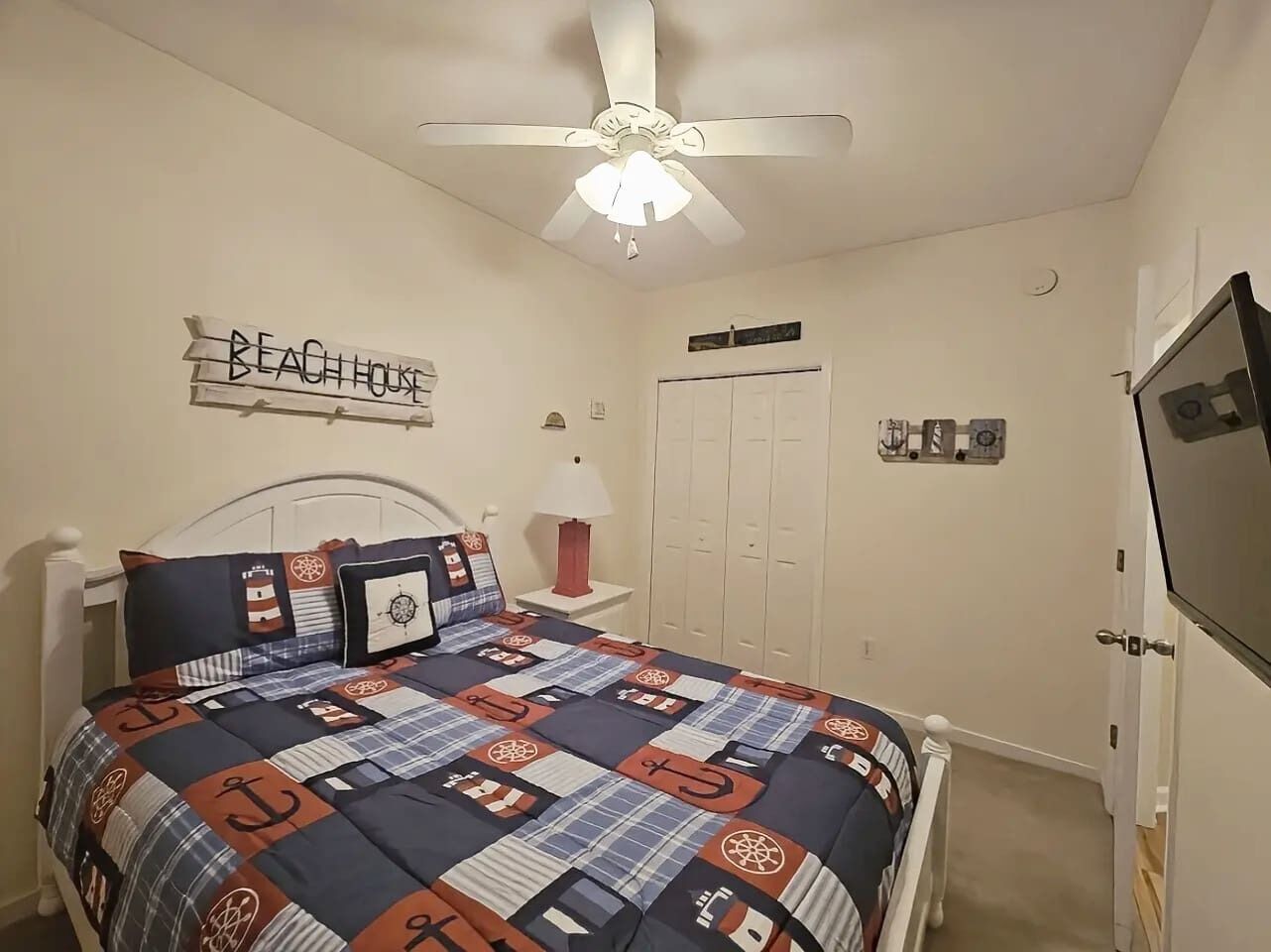 A bedroom with a bed, a ceiling fan, a TV, a wardrobe, and a wall sign that reads 'BEACH HOUSE'.