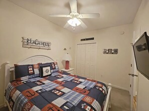 Ein Schlafzimmer mit Bett, Deckenventilator, Fernseher, Kleiderschrank und einer Wandtafel mit der Aufschrift 'BEACH HOUSE'.