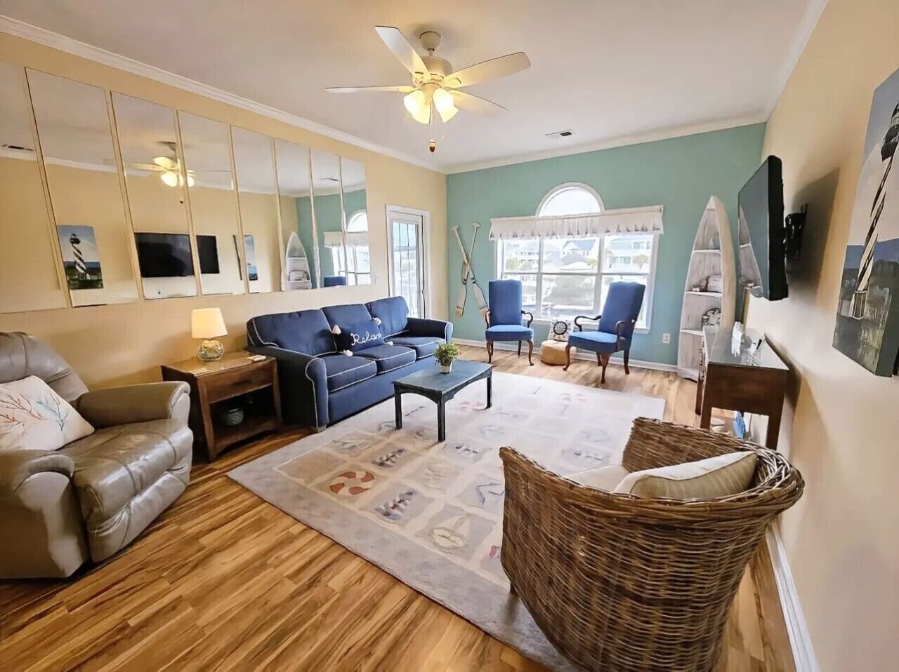 A living room with a sofa, armchair, and a wicker chair. There is a ceiling fan, a television, and a window with curtains.